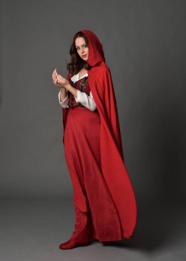 Uzun etekli ve dalgalı kapüşonlu pelerinli kırmızı ortaçağ fantezi kostümü giyen güzel esmer kadının tam boy portresi. El hareketleriyle poz veriyor, gri stüdyo arka planında izole edilmiş..