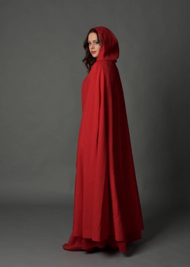 Kırmızı ortaçağ fantezi kostümü giymiş, kapüşonlu pelerinli bir kadının uzun bir portresi. Arka planda duran, el hareketleriyle poz veren, gri stüdyo arkaplanındaki kameradan uzaklaşan....