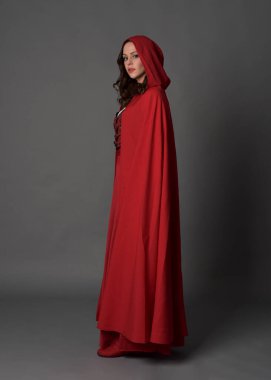 Kırmızı ortaçağ fantezi kostümü giymiş, kapüşonlu pelerinli bir kadının uzun bir portresi. Duruş profili, hareketli el pozları, gri stüdyo arka planında izole edilmiş kameradan uzaklaşmak..