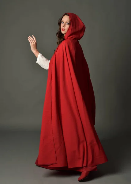 stock image Full length portrait of woman wearing red medieval fantasy costume, flowing hooded cloak. Standing pose in backview, gestural hand poses, walking away from camera isolated on grey studio background.