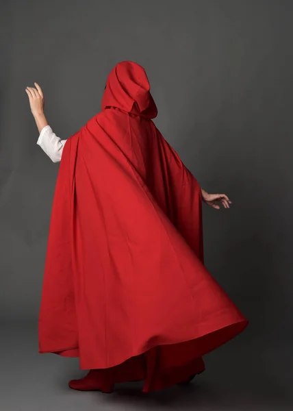赤い中世のファンタジーの衣装を着て女性の完全な長さの肖像画 フード付きのマントを流れる 背景色の中に立つポーズ ジェスチャーの手ポーズ 灰色のスタジオの背景に隔離されたカメラから離れて歩く — ストック写真