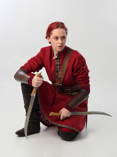  portrait of beautiful woman wearing a red medieval fantasy warrior costume with leather armour, holding weapons.  Standing pose isolated on studio background.