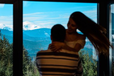 Adam kızı kollarıyla pencerenin arka planında, güzel bir dağ manzarasının arka planında, Karpatlar 'da bir evde tutuyor..