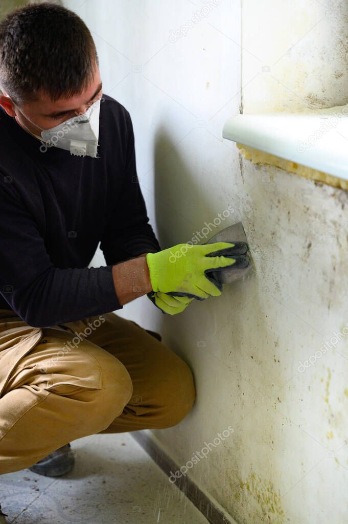 A man with a spatula removes mold and fungus on the wall, a man in a mask and gloves.