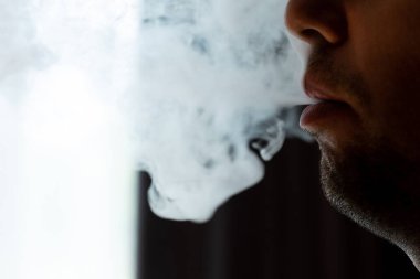 Male lips blowing smoke close-up, cigarette smoke and lips on black background, male unshaven and short beard.