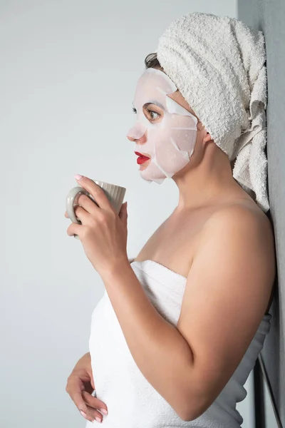 Facial care, a girl and a fabric mask on her face, a woman holds a cup with a drink in her hands.