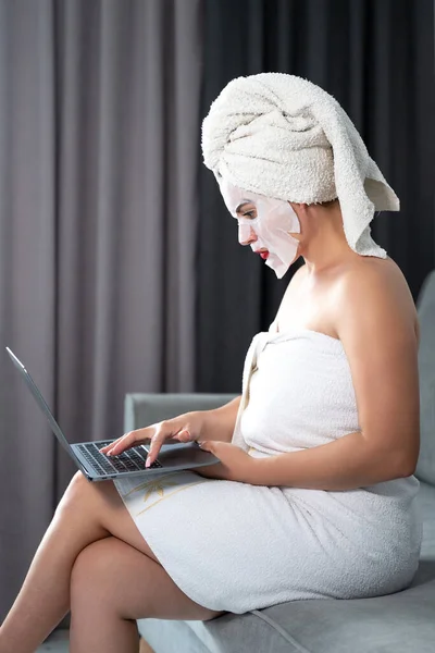 A woman during a cosmetic procedure works with a laptop, remote work mode, cosmetic procedures with a face, a girl in a towel.