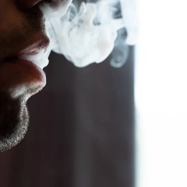 Seductive male lips blowing cigarette smoke, light stubble on beard, portrait of male lips and smoke on black background.