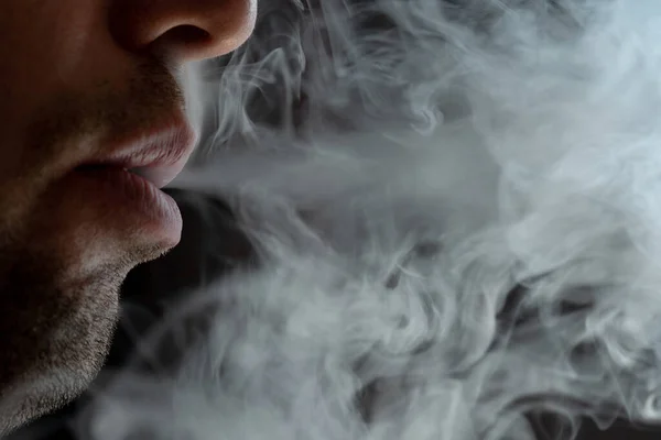 Seductive male lips blowing cigarette smoke, light stubble on beard, portrait of male lips and smoke on black background.