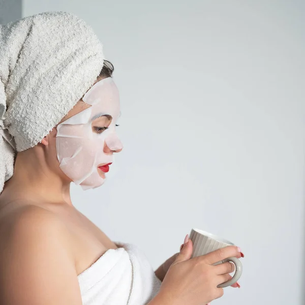 Facial care, a girl and a fabric mask on her face, a woman holds a cup with a drink in her hands.