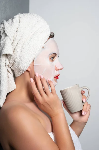 Fabric face mask, moisturizing mask for facial skin, girl holding a cup in her hands.