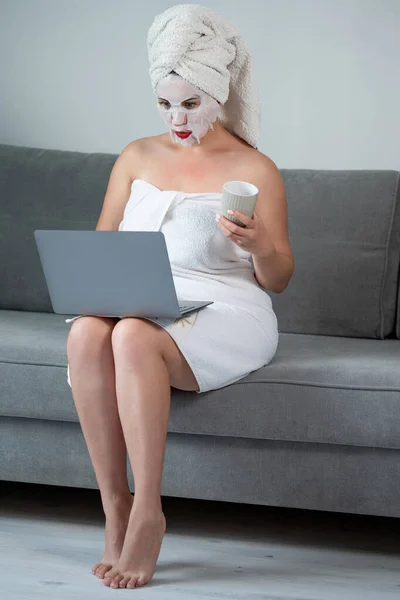A woman during a cosmetic procedure works with a laptop, remote work mode, cosmetic procedures with a face, a girl in a towel.