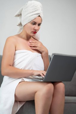 Woman in towel sitting on sofa and working remotely online with laptop, work from home with rest, wireless connection.