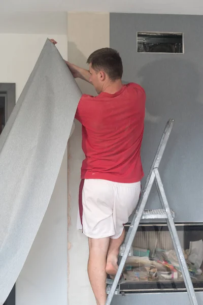 A man glues wallpaper on a wall in a house, a craftsman uses a ladder to glue wallpaper on top.