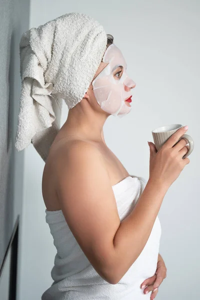 Facial care, a girl and a fabric mask on her face, a woman holds a cup with a drink in her hands.