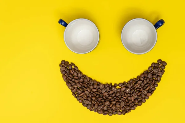 Happy emoticon made of coffee beans on a yellow background, Smiley made of coffee and two empty cups as eyes.