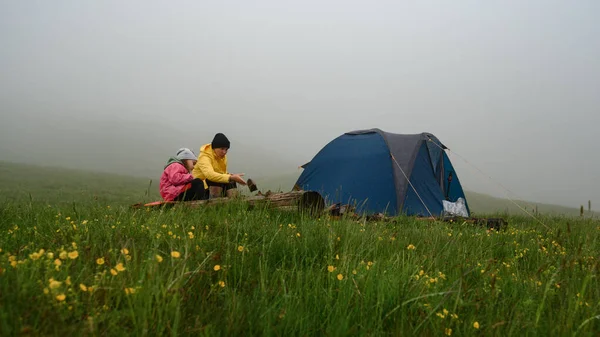 Mother and daughter sit near the tent after the rain, family vacation in the mountains, fog in the mountains.