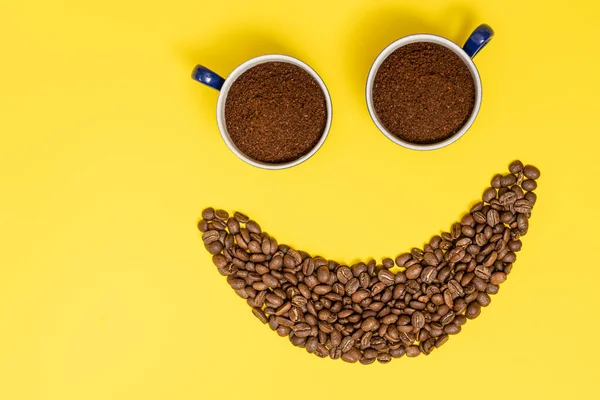Happy emoticon made of coffee beans on a yellow background, Smiley made of coffee and two cups of ground coffee as eyes.