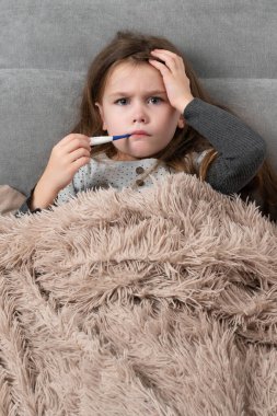 A cute little girl is lying in bed sick, the child measures the temperature with a children's electronic thermometer.