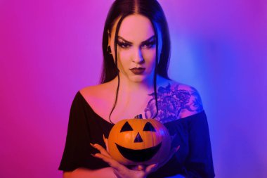 Muhteşem esmer cadı elinde bir balkabağı tutuyor ve kameraya bakıyor. Korkunç Cadılar Bayramı fotoğrafı. Kız Cadılar Bayramı 'nı neon ışıkla kutluyor.