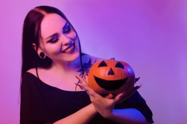 Muhteşem esmer cadı elinde bir balkabağı tutuyor ve kameraya bakıyor. Korkunç Cadılar Bayramı fotoğrafı. Kız Cadılar Bayramı 'nı neon ışıkla kutluyor.