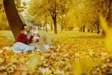 Aşka düşen sonbahar yaprakları bir parkta oturan çift, bir ağacın yanında güzel bir sonbahar günü zevk otur. Mutlu neşeli genç Aile Baba, anne ve bebek kız açık havada eğleniyor