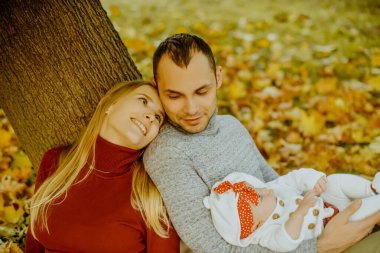 Aşka düşen sonbahar yaprakları bir parkta oturan çift, bir ağacın yanında güzel bir sonbahar günü zevk otur. Mutlu neşeli genç Aile Baba, anne ve bebek kız açık havada eğleniyor