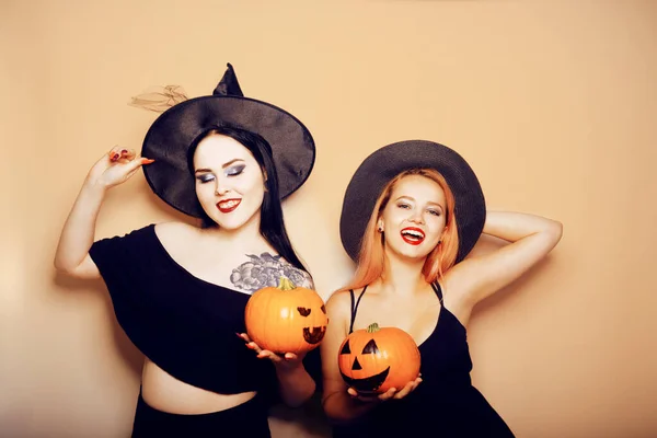Two girls witches in hats are holding pumpkins in their hands and smiling for the camera, posing, dancing. Women celebrate Halloween, photo on a red background