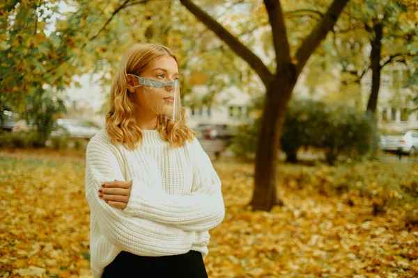 Sonbaharda parkta güzel bir kız. Şeffaf maskeli bir kadın caddede yürüyor. Covid tarzı. Maskeli genç.