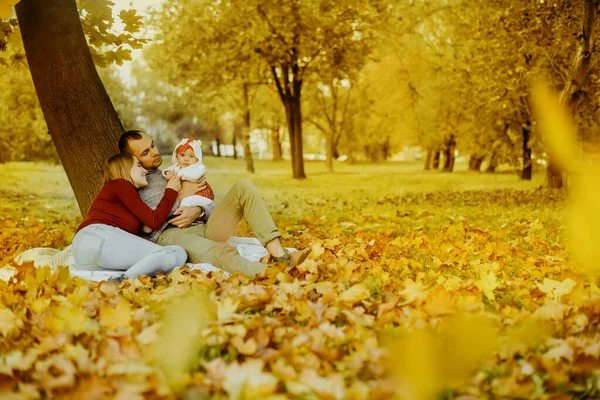 Aşka düşen sonbahar yaprakları bir parkta oturan çift, bir ağacın yanında güzel bir sonbahar günü zevk otur. Mutlu neşeli genç Aile Baba, anne ve bebek kız açık havada eğleniyor