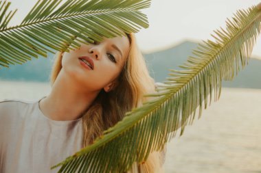 Güzel, genç bir sarışın, elinde palmiye dalları tutuyor. Temiz ve güzel bir cilt, yüz. Dağların ve denizin arka planında bir kız