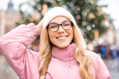 Noel ağacının arka planında gözlüklü bir kızın şirin portresi. Kış, ilk kar, pembe ceketli bir sarışın ve beyaz şapkalı bir gülümseme. Yeni Yıl