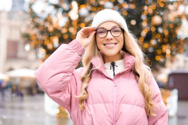 Noel ağacının arka planında gözlüklü bir kızın şirin portresi. Kış, ilk kar, pembe ceketli bir sarışın ve beyaz şapkalı bir gülümseme. Yeni Yıl