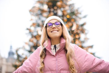 Noel ağacının arka planında gözlüklü bir kızın şirin portresi. Kış, ilk kar, pembe ceketli bir sarışın ve beyaz şapkalı bir gülümseme. Yeni Yıl