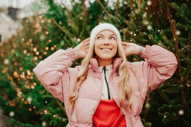 Şapkalı ve pembe ceketli hoş bir sarışın Noel ağaçlarının arkasında duruyor. Bir kız Noel ağacı alır, yeni yıl havası, kış, tatiller, yeni yıl gülümsemesi
