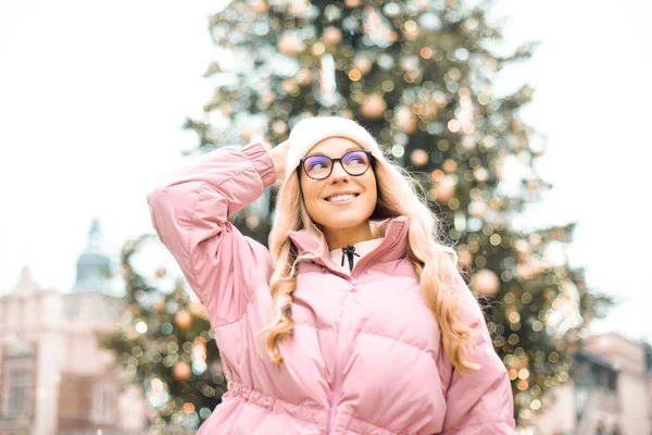 Noel ağacının arka planında gözlüklü bir kızın şirin portresi. Kış, ilk kar, pembe ceketli bir sarışın ve beyaz şapkalı bir gülümseme. Yeni Yıl