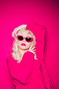 Sarışın, pembe bir arka planda şık pembe gözlüklü ve elbiseli poz veriyor. Kız model pozu, parlak fotoğraf tarzı, altın mücevher 80 'ler ve 90' lar..