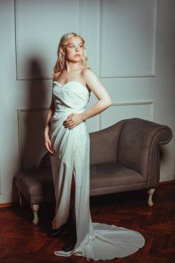 Gelin duvarın önünde duruyor, şık bir elbise. Gelinlik salonu. Beyaz, uzun küpeli bir kadın. Büyümüş güzel moda sarışın. Siyah çizmeler.