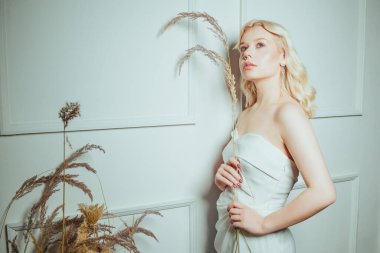 Gelin duvara yaslanır ve düğün için seçeceği çiçekleri tutan bir çivi tutar. Beyaz elbiseli, uzun küpeli bir kadın. Güzel sarışın.