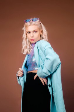 Turkuaz ceketli, pullu mavi ceketli bir sarışın stüdyoda kahverengi arka planda poz veriyor. Yüksek moda, 90 'ların 80' leri. Kız dans ediyor ve hareket ediyor. Pembe makyajlı bir model.