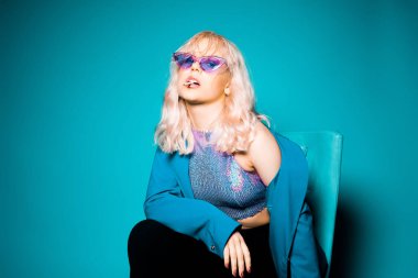 Modaya uygun, mavi ceketli, gözlüklü, 90 'lı yılların modasına uygun sarışın bir kadın koltuğa oturup poz veriyor. Mavi arka planda. Modaya uygun bir kadın sigara içiyor..