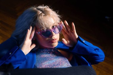 Bu kız 80 'lerden kalma klipteki gibi. Kısa sarı saç kesimi. Kız mavi ceketli, parlak ceketli ve gözlüklü bir sandalyede oturuyor. Moda & Biçimi