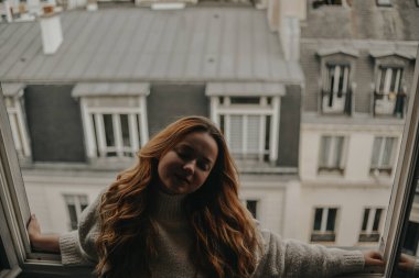Bir kadın pencerenin yanında duruyor Paris 'in çatılarının arka planına karşı. Bej takım elbiseli, uzun saçlı kız..