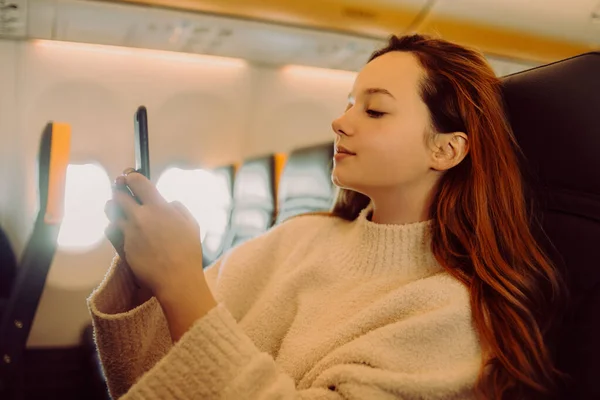 一个漂亮的女孩坐在飞机上看着电话 看书或玩游戏 空中旅行 长途飞行 — 图库照片