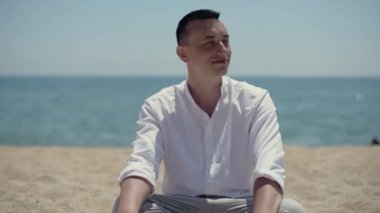 Bir adam denizin ya da okyanusun arka planına karşı sahilde oturur ve kameraya bakar. Beyaz gömlekli adam gülümsüyor..