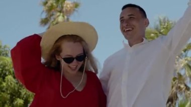 A fashionable couple is dancing against the backdrop of palm trees. A girl in a red dress, glasses and a hat, a man in a white shirt. Vacation, vacation, summer.