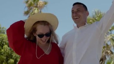 A fashionable couple is dancing against the backdrop of palm trees. A girl in a red dress, glasses and a hat, a man in a white shirt. Vacation, vacation, summer.
