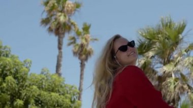 Happy girl in a red dress on vacation on the background of palm trees laughing and smiling. Vacation. A woman in sunglasses and a big hat enjoys life and dances.