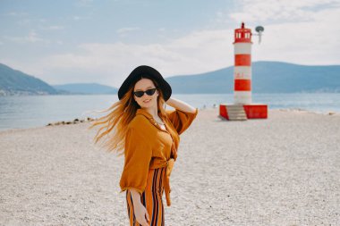 Sahilde sarı-turuncu takım elbiseli bir kız denizin arka planında, dağlarda ve deniz fenerinde. Güneş gözlüklü ve şapka danslı moda kadını.