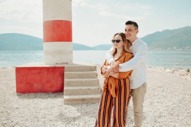 Sahildeki deniz fenerinin yanında, denizin ve dağların arkasında bir çift duruyor. Tatil aşkı, balayı, seyahat. Tarz sahibi çift gülüyor ve gülüyor